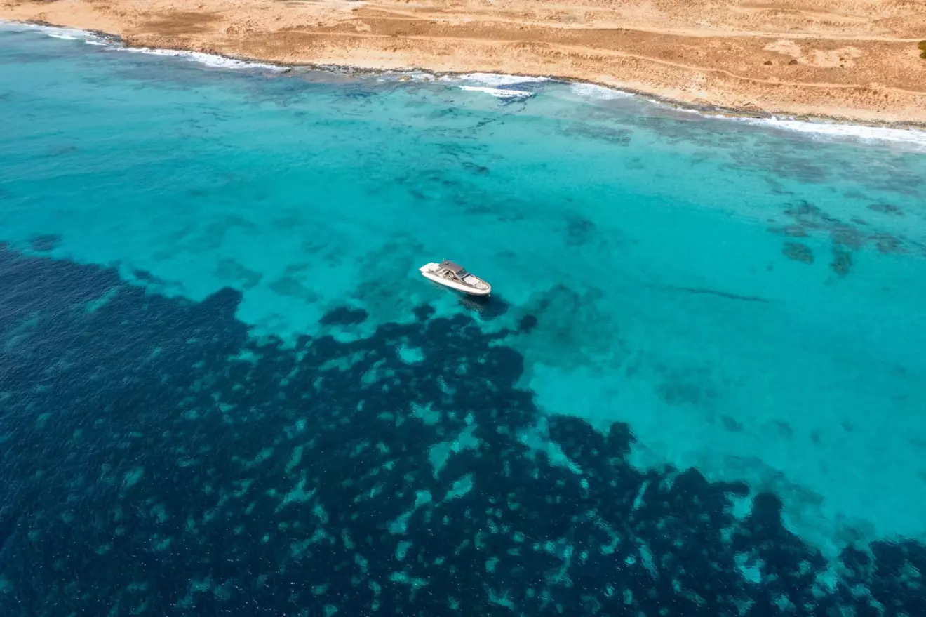 Aerial view of a SACS Rebel 47 white boat anchored in crystal clear turquoise water near a sandy beach.